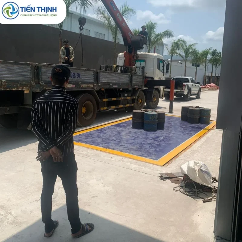 Cân hàng hóa với các thùng chứa, có xe tải và người đang điều khiển cần cẩu.